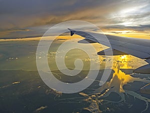 Airplane wing flying over sea and estuary at sunset time. Orange light reflect with ocean. Travel concept.