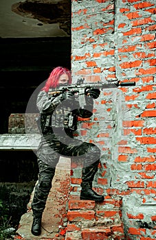 Airsoft red-hair woman in uniform with machine gun beside brick wall. Soldier aims at the sight on the ruins. Vertical photo side photo