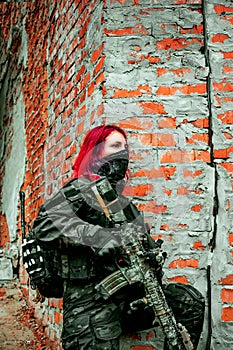 Airsoft red-hair woman in uniform with machine gun standing beside brick wall. Soldier on ruine. Vertical photo