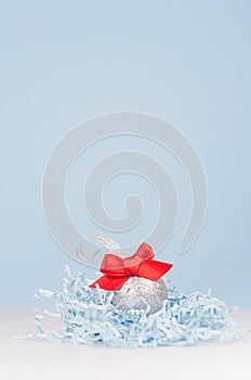 Airy soft light christmas minimalist background - silver small ball with red bow on white and pastel blue backdrop, copy space.