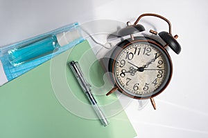 Alarm clock along with note blog and fountain pen, mask and disinfectant,green paper, on gray background