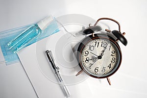 Alarm clock along with note blog and fountain pen, mask and disinfectant,white paper,on gray background