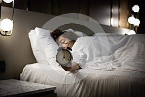 Alarm clock on the bed with sleeping woman behind. Girl keeps alarm clock by hand