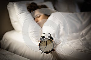 Alarm clock on the bed with sleeping woman behind. Girl keeps alarm clock by hand