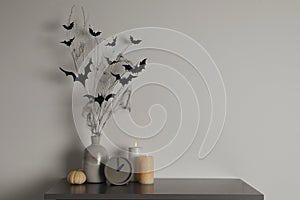 Alarm clock, burning candles and vase with paper bats  on table near white wall, space for text. Halloween decor