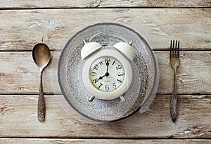 Alarm clock on ceramic plate, vintage spoon and fork on old wooden background