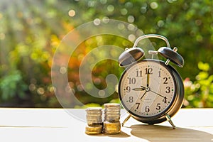 Alarm clock and coins on the wooden table in the sunset, money concept.