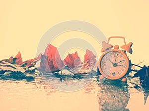 Alarm clock in colorful autumn leaves against a dark background with shallow depth of field. Daylight savings time concept. Alarm