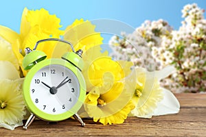 Alarm clock and flowers on wooden table against blurred background. Spring time