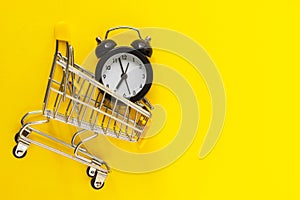 Alarm clock in mini shopping cart on yellow background. Buying, time, sale concept. Flat lay, top view