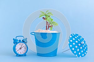 Alarm clock, paper cocktail parasol, can with sand and plastic palm tree isolated on blue background. Summertime vacation minimali