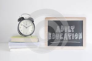 Alarm clock on a pile of books next to a blackboard with the text ADULT EDUCATION wrote on it. Empty copy space