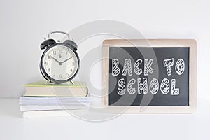 Alarm clock on a pile of books next to a blackboard with the text BACK TO SHOOL wrote on it. Empty copy space