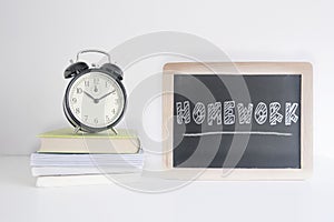 Alarm clock on a pile of books next to a blackboard with the text HOMEWORK wrote on it. Empty copy space