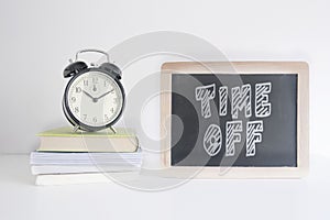 Alarm clock on a pile of books next to a blackboard with the text TIME OFF wrote on it. Empty copy space