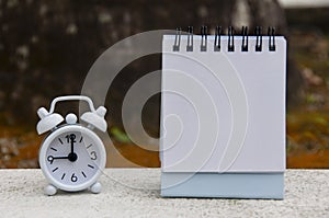 Alarm clock pointing at 9 o'clock with blank notepad. Blurred park background. Copy space concept