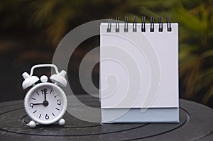 Alarm clock pointing at 9 o'clock with empty notepad. Blurred park background. Copy space concept