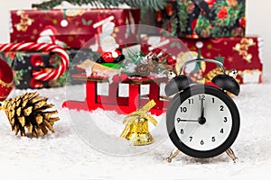 Alarm clock, santa claus sleigh, boxes with gifts, christmas tree cone and golden bell on a background of snow