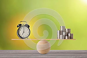 Alarm clock on seesaw balancing with stacking coins money on wooden table, meaning of business investment and saving growth with