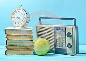 Alarm clock on stack of old books, radio receiver, apple on a blue background. Retro still life.
