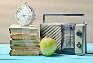 Alarm clock on stack of old books, radio receiver, apple on a blue wooden table. Retro still life.