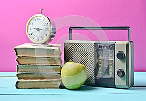 Alarm clock on stack of old books, radio receiver, apple on a pink background. Retro still life.