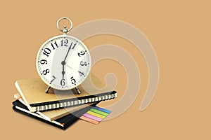 Alarm clock stacked on brown and black notebook with a post it note paper. Isolated on brown background with copy space and clip.