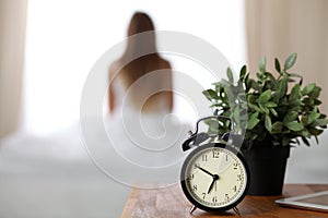 Alarm clock standing on bedside table has already rung early morning to wake up woman in bed sitting in background
