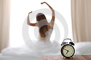 Alarm clock standing on bedside table has already rung early morning to wake up woman in bed sitting in background