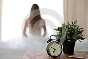 Alarm clock standing on bedside table has already rung early morning to wake up woman in bed sitting in background