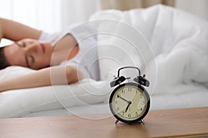 Alarm clock standing on bedside table has already rung early morning to wake up woman in bed sleeping in background