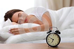 Alarm clock standing on bedside table has already rung early morning to wake up woman in bed sleeping in background