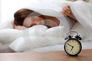 Alarm clock standing on bedside table has already rung early morning to wake up woman in bed sleeping in background