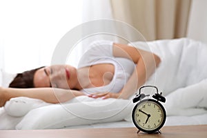 Alarm clock standing on bedside table has already rung early morning to wake up woman in bed sleeping in background