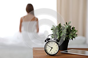 Alarm clock standing on bedside table has already rung early morning to wake up woman is stretching in bed in background
