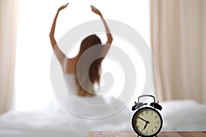 Alarm clock standing on bedside table has already rung early morning to wake up woman is stretching in bed in background