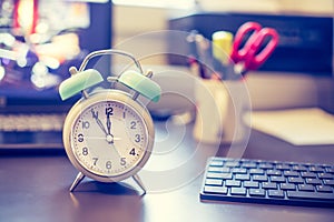 Alarm clock is standing on a grey desk, office stuff in the blurry background