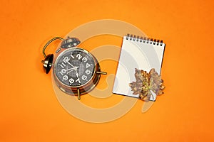 Alarm o`clock showing 7 o`clock and autumn leaves and empty notebook  isolated on orange background