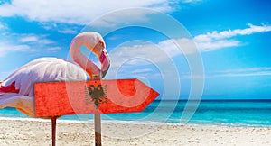 Albania flag on wooden table sign on beach background with pink flamingo. There is beach and clear water of sea and blue sky in