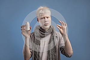 Albino guy holding jar with meds and pill in hands, standing on blue background, taking anti viral medication