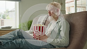 Albino man watching scary movie, sitting on sofa in large room, holding popcorn