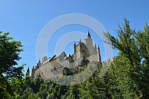 Alcazar Castle Seen From The River That Runs Through The Valley That Reigns In Segovia Slightly Caught By A Small Grove. Architect