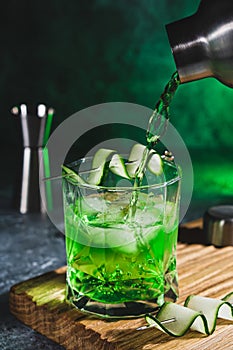 Alcohol cocktail with cucumber pouring from shaker in whiskey glass with ice cubes. Summer coctail
