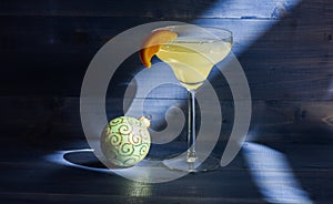 Alcohol cocktail margarita with piece of orange near christmas ball ornament on dark blue background. Celebrate new year