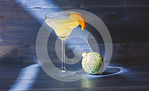 Alcohol cocktail margarita with piece of orange near christmas ball ornament on dark blue background. Celebrate new year