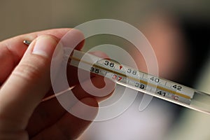 An alcohol thermometer shows a temperature of 37.9 on a blurred background of a sick girl lying on a bed. Check the temperature.