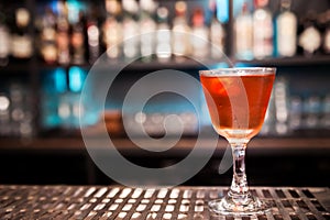 An alcoholic cocktail of red color with a decoration of lemon peel stands on a bar counter.