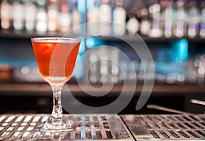 An alcoholic cocktail of red color with a decoration of lemon peel stands on a bar counter.