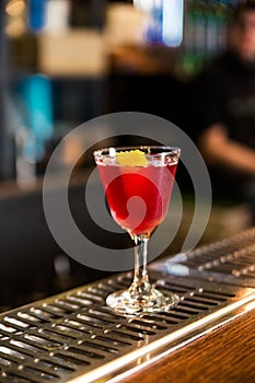 An alcoholic cocktail of red color with a decoration of lemon peel stands on a bar counter