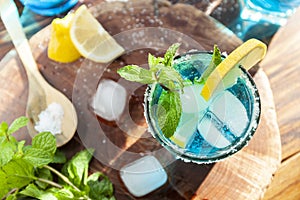 Alcoholic cocktail on a wooden table surrounded by ingredients such as lemon, salt, sugar, ice